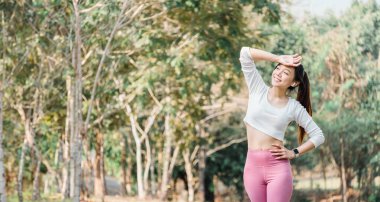 Beyaz elbiseli neşeli bir kadın ve alnını silen pembe taytlar, arka planda hafifçe odaklanmış ağaçlar..