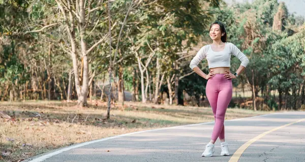 Smiling woman in a white cropped top and pink leggings stands with hands on hips on a paved park pathway, trees in the background.