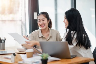 İki gülümseyen iş kadını çalışma masasında laptopları ve kahve fincanlarıyla belgeleri paylaşıyor..