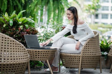 Nişanlı serbest çalışan biri dizüstü bilgisayarı üzerinde çalışmak için öne doğru eğiliyor, etrafı yemyeşil bitkilerle dolu, davetkar bir bahçede oturuyor..