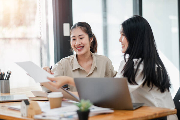Две улыбающиеся деловые женщины обмениваются документами за рабочим столом с ноутбуками и кофейными чашками.