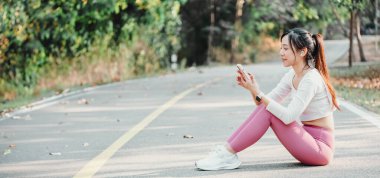 Pembe taytlı ve beyaz spor ayakkabılı kadın akıllı telefonunu park yolunda, kulaklıkları ve arka planda ağaçlarıyla kullanıyor..