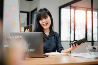Neşeli iş kadını, arka planda yapışkan notlarla dolu bir ofisteki ahşap masasında hazır bir dizüstü bilgisayarla, akıllı telefonuyla bir sohbete girişiyor..
