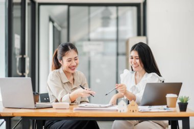İki kadın bir masada dizüstü bilgisayarlar ve kalemlerle oturuyor, gülümsüyor ve konuşuyorlar. Sahne dostane ve işbirlikçidir.