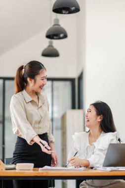 Ayakta duran kadın, masa başında dizüstü bilgisayarları ve çalışma materyalleri olan modern bir ofis ortamında iş tartışırken, oturan meslektaşına gülümsüyor..
