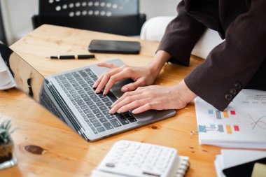 Bir kadın hesap makinesi, cep telefonu ve fincanı olan bir masanın önünde dizüstü bilgisayar kullanıyor.