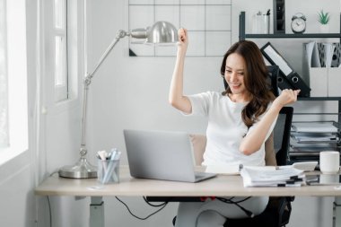 Bir kadın elinde dizüstü bilgisayar ve bir fincan kahveyle masada oturuyor. Gülümsüyor ve mutlu.
