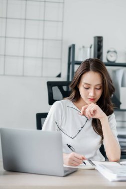 Özenli genç iş kadını işine odaklanıyor, modern ofis ortamındaki bir masada dizüstü bilgisayarla belgelere bakıyor..