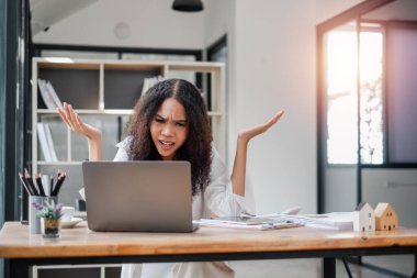 Kafası karışmış iş kadını, belgelerle çevrili modern bir ofis masasında dizüstü bilgisayarıyla çalışırken hayal kırıklığı yaşıyor..