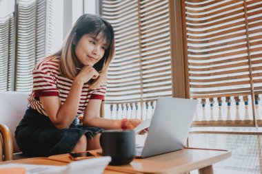 Genç Asyalı kadın, doğal ışık alan rahat bir kafede dizüstü bilgisayarıyla dikkatlice çalışıyor..