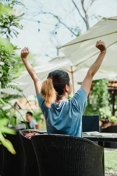 A remote worker stretches with raised arms in triumph in front of a laptop at an outdoor home office setting.