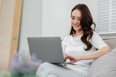 Mutlu bir kadın evdeki bir kanepede dizüstü bilgisayar kullanarak rahatça vakit geçirir ya da rahatça çalışır..