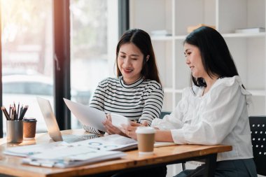 Bir proje üzerinde çalışan iki meşgul bayan meslektaşı, iyi aydınlatılmış bir ofis alanında dokümanlar üzerine fikir alışverişinde bulunuyorlar..