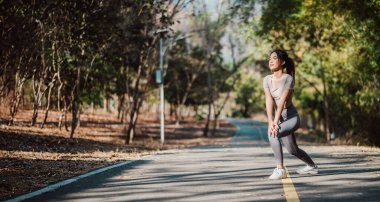 Fitness bilinci olan bir kadın ağaçlarla çevrili güneşli bir yolda esneyerek sabah koşusuna hazırlanıyor..