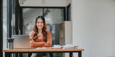 Hoş bir gülümsemesi olan profesyonel bir genç kadın iyi organize edilmiş bir ofiste dizüstü bilgisayarıyla çalışıyor..