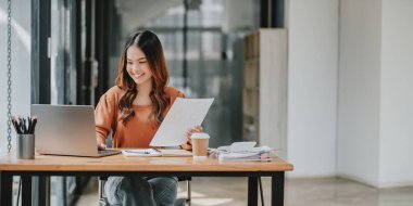 Genç iş kadını bir belgeyi incelerken gülümsüyor, geniş bir ofiste laptopuyla rahatça çalışıyor..