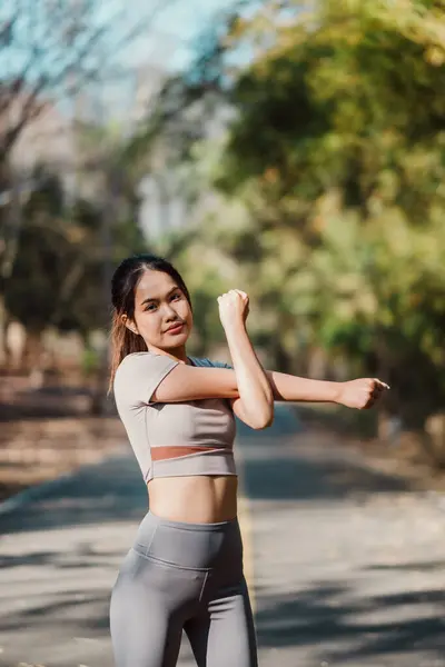 Motivated young woman in fitness attire stretching her arms, preparing for a workout on a park path surrounded by trees.