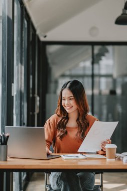 Genç bir kadın, güneşli ve çağdaş bir ofis alanında dizüstü bilgisayarla çalışırken gülümsüyor..