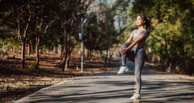 Atletik kadın, ağaçlar ve doğa ile çevrili sakin bir patikada ayakta durarak bacaklarını esnetiyor..