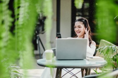 Dizüstü bilgisayarında çalışan neşeli bir kadın ve açık bir kafede akıllı telefon kullanıyor. Etrafı yemyeşil bitkilerle çevrili..