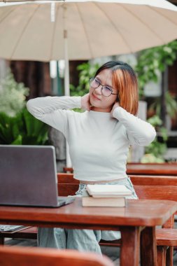 Genç bir kadın, dışarıda tahta bir masada laptopunun başında dinlenmek için boynunu biraz uzatıyor..