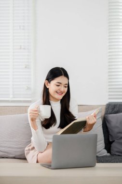 Genç bir kadın, önünde dizüstü bilgisayarı açık bir şekilde kitap okurken kahve içmekten zevk alıyor. Bu da iş hayatının sakin bir denge içinde olduğunu gösteriyor..