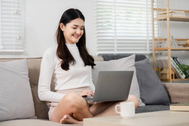 Neşeli genç bir kadın rahat bir şekilde koltukta oturur, parlak ve rahat bir ev ortamında dizüstü bilgisayarla çalışır, esnek bir uzaktan kumandanın örneğini verir..