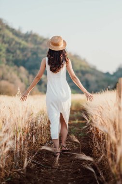 Beyaz elbiseli ve hasır şapkalı bir kadın buğday tarlasında zarif bir şekilde yürüyor. Doğayla bir bağ ve özgürlük hissi uyandırıyor..