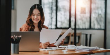 Bir kadın elinde dizüstü bilgisayar ve bir yığın kağıtla masada oturuyor. Gülümsüyor ve işinden zevk alıyor.