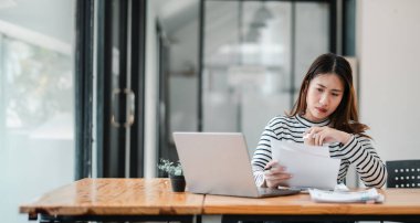 Bir kadın elinde dizüstü bilgisayar ve bir yığın kağıtla masada oturuyor. Gazetelere bakıyor ve ciddi ya da odaklanmış bir ruh halinde.