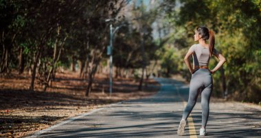 Arka planda ağaçlar olan bir kadın yolda koşuyor. Gri bir gömlek ve gri pantolon giyiyor.