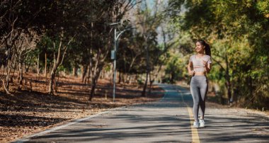 Arka planda ağaçlar olan bir kadın yolda koşuyor. O, pembe bir gömlek ve gri pantolon giyiyor.