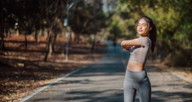 Bir kadın yolda geriniyor. Gri bir gömlek ve gri pantolon giyiyor. Yol boş ve güneş parlıyor.