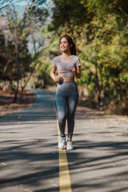 Arka planda ağaçlar olan bir kadın yolda koşuyor. Gri bir gömlek ve gri pantolon giyiyor.