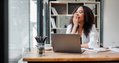 Bir kadın masasında dizüstü bilgisayarının üzerinde uyuyor. O, beyaz bir gömlek giyiyor ve masasında bir saksı çiçeği var.