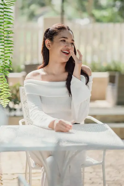 stock image A woman is sitting at a table with a smile on her face. She is wearing a white shirt and is talking on her cell phone. The scene is set outdoors, with a bench and a potted plant nearby