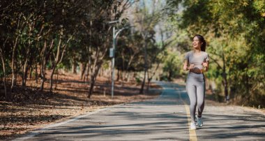 Arka planda ağaçlar olan bir kadın yolda koşuyor. Gri bir gömlek ve gri pantolon giyiyor.