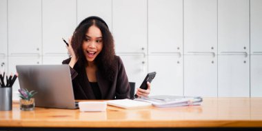 Bir kadın dizüstü bilgisayarı ve cep telefonu olan bir masada oturuyor. Gülümsüyor ve mutlu.