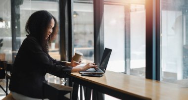 Bir kadın elinde dizüstü bilgisayar ve bir fincan kahveyle masada oturuyor. O, işine odaklanmış ve ciddi bir ruh hali içinde.