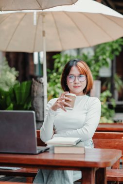 Bir kadın elinde dizüstü bilgisayar ve bir fincan kahveyle masada oturuyor. O, gözlük takıyor ve yüzünde bir gülümseme var. Sahne rahat ve rahat bir atmosferi gösteriyor.