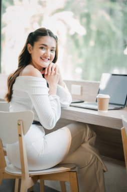 Masasında dizüstü bilgisayarı ve bir fincan kahvesiyle oturan bir kadın. Gülümsüyor ve eğleniyor.