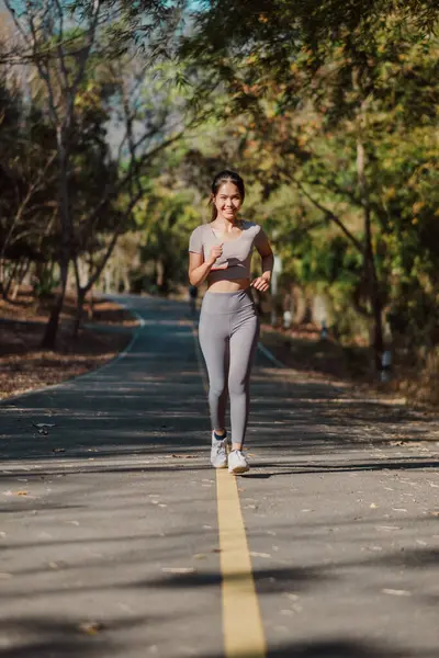 A woman is running on a path in a park. She is wearing a grey shirt and grey pants