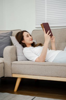 Bir kadın kanepeye uzanmış kitap okuyor. Gülümsüyor ve eğleniyor.