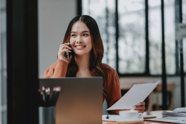 Bir kadın cep telefonuyla konuşuyor bir masada dizüstü bilgisayar ve fincanla otururken. Gülümsüyor ve sohbetten zevk alıyor.