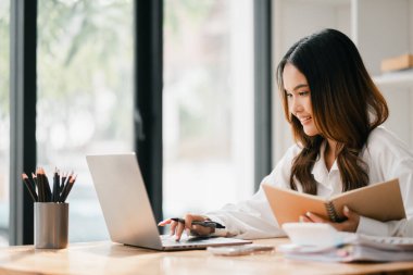 Masasında dizüstü bilgisayarı ve not defteri olan bir kadın oturuyor. Gülümsüyor ve işinden zevk alıyor.