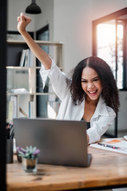 Bir kadın dizüstü bilgisayarı ve saksısı olan bir masada oturuyor. Gülümsüyor ve mutlu.