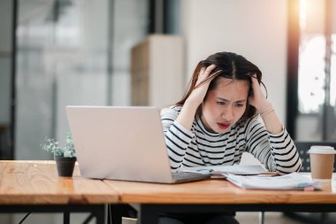 A woman is sitting at a desk with a laptop and a cup of coffee. She is looking at the laptop with a worried expression on her face