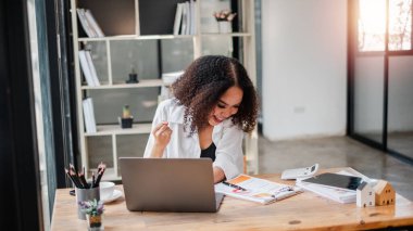 Bir kadın elinde dizüstü bilgisayar ve bir yığın kağıtla masada oturuyor. Gülümsüyor ve mutlu.