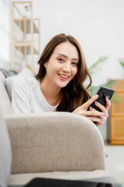 A woman is sitting on a couch and smiling while holding a cell phone. Concept of relaxation and leisure, as the woman is enjoying her time on the couch