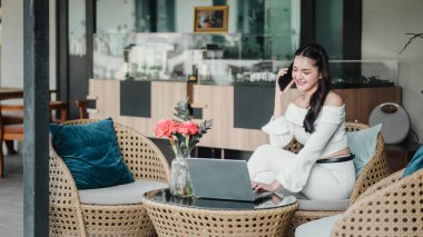 Bir kadın bir dizüstü bilgisayar ve bir vazo çiçekle bir masada oturuyor. O, cep telefonuyla konuşuyor. Sahne rahat ve rahat, kadın dışarıda eğleniyor.